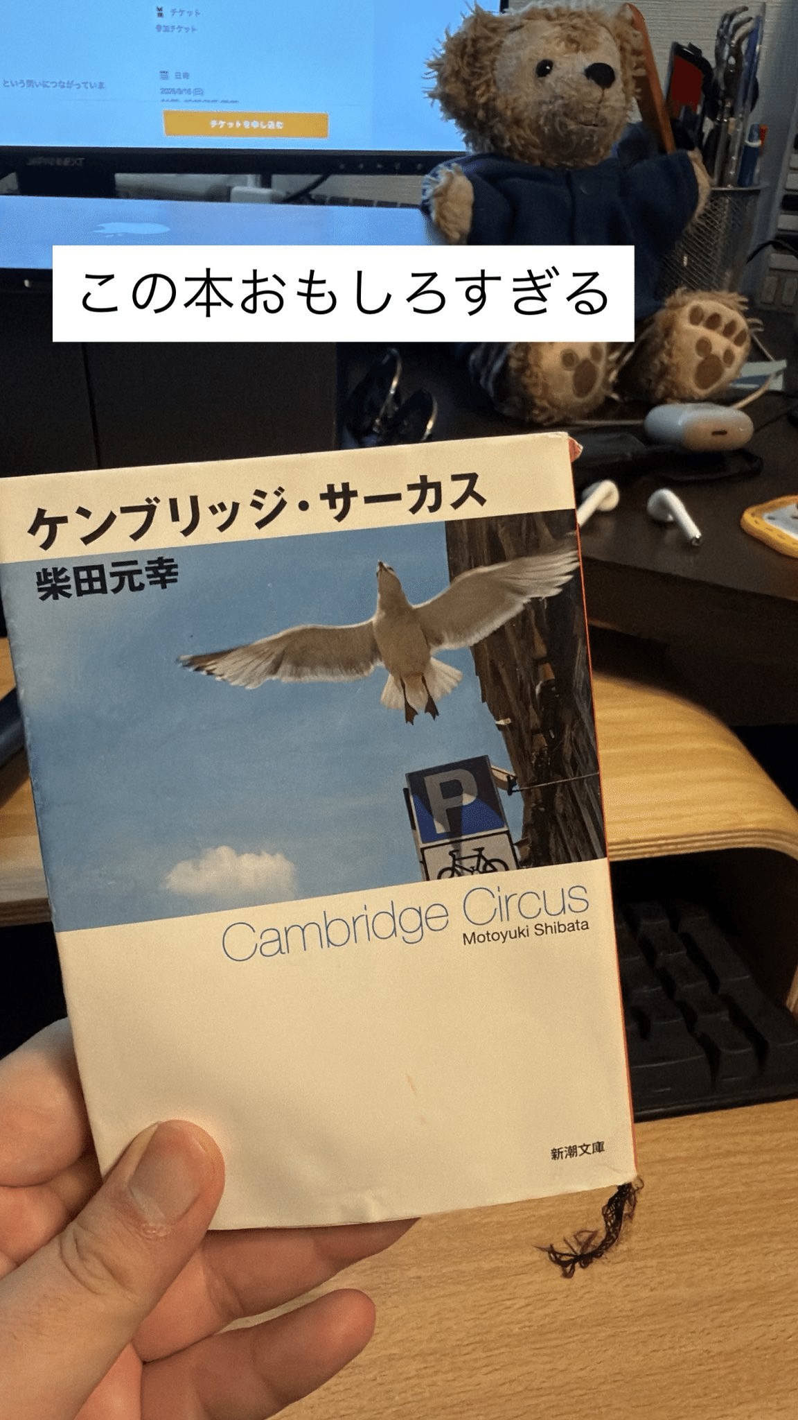 ALT: 左手が持っている、柴田元幸著『ケンブリッジ・サーカス』の新潮文庫本の表紙。表紙には、日本語で「ケンブリッジ・サーカス」と著者名、英語で「Cambridge Circus Motoyuki Shibata」のタイトル、そして青空を飛ぶカモメの画像が描かれている。本の背後に浮かぶ白いテキストボックスには、日本語で「この本おもしろすぎる」と書かれている。背景には、木目調のデスクの上にキーボード、ワイヤレスイヤホン、眼鏡、青いセーターを着たテディベアのぬいぐるみ、そしてブラウザ画面が表示されたPCモニターが見える。