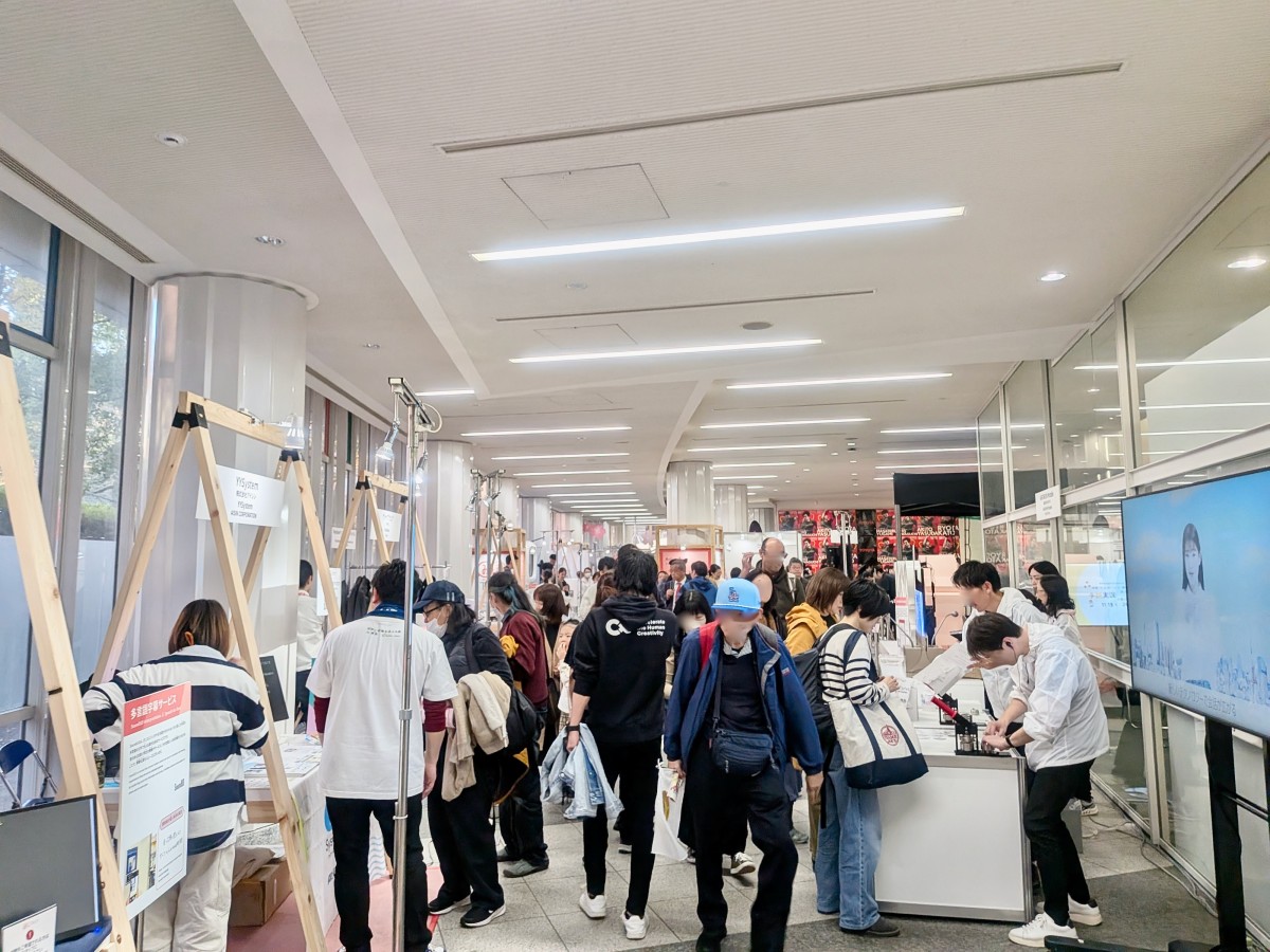 施設内の屋内技術展示エリア。企業や役所のような白い天井と廊下に、いろいろな企業・団体が出展している。見学者でにぎわっており熱心に見ている