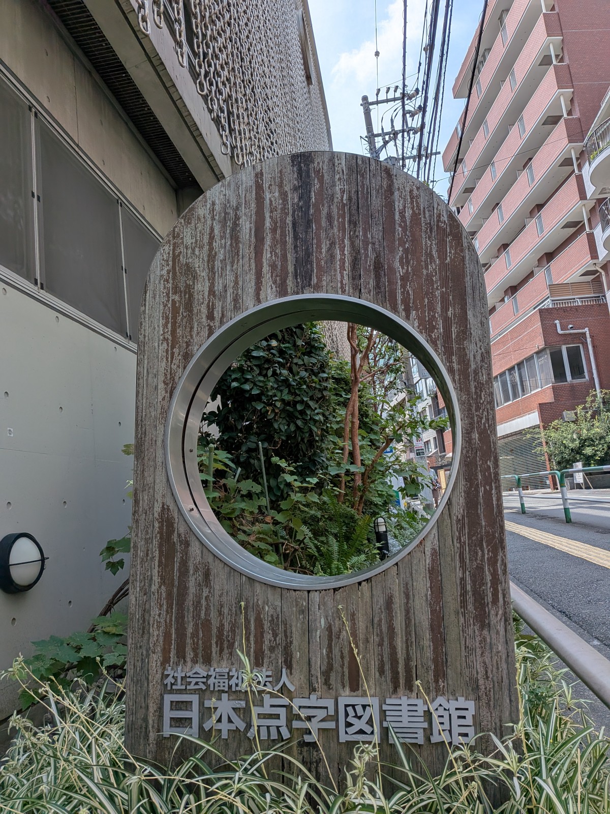 日本点字図書館のエントランスにある看板。木の板に大きな穴があいており、その下に日本点字図書館と銘記されている。