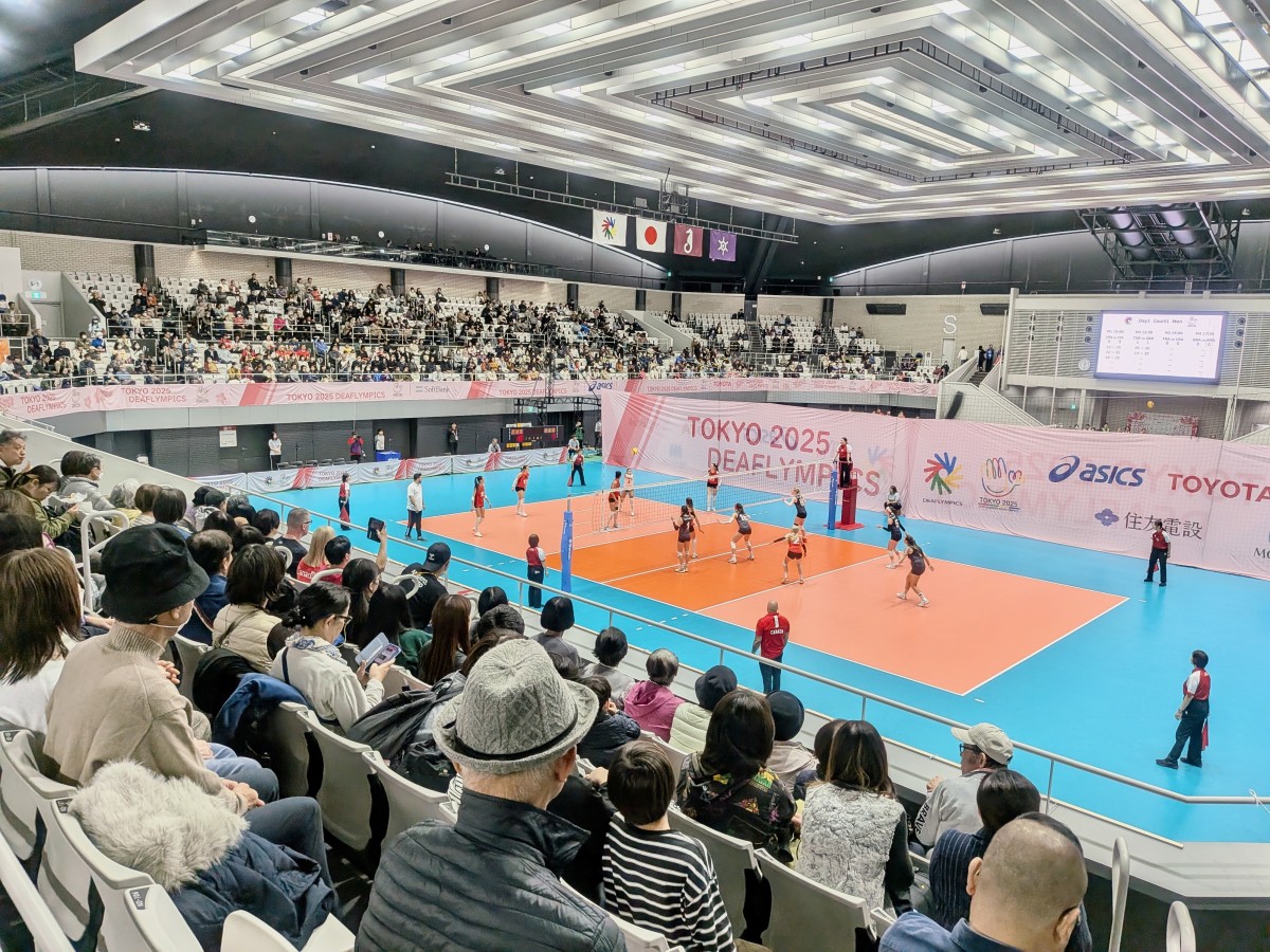 試合中のコート。直線的なデザインの未来感のある照明の下、女子選手たちがプレーしている。ブルーのフロアにオレンジのコート