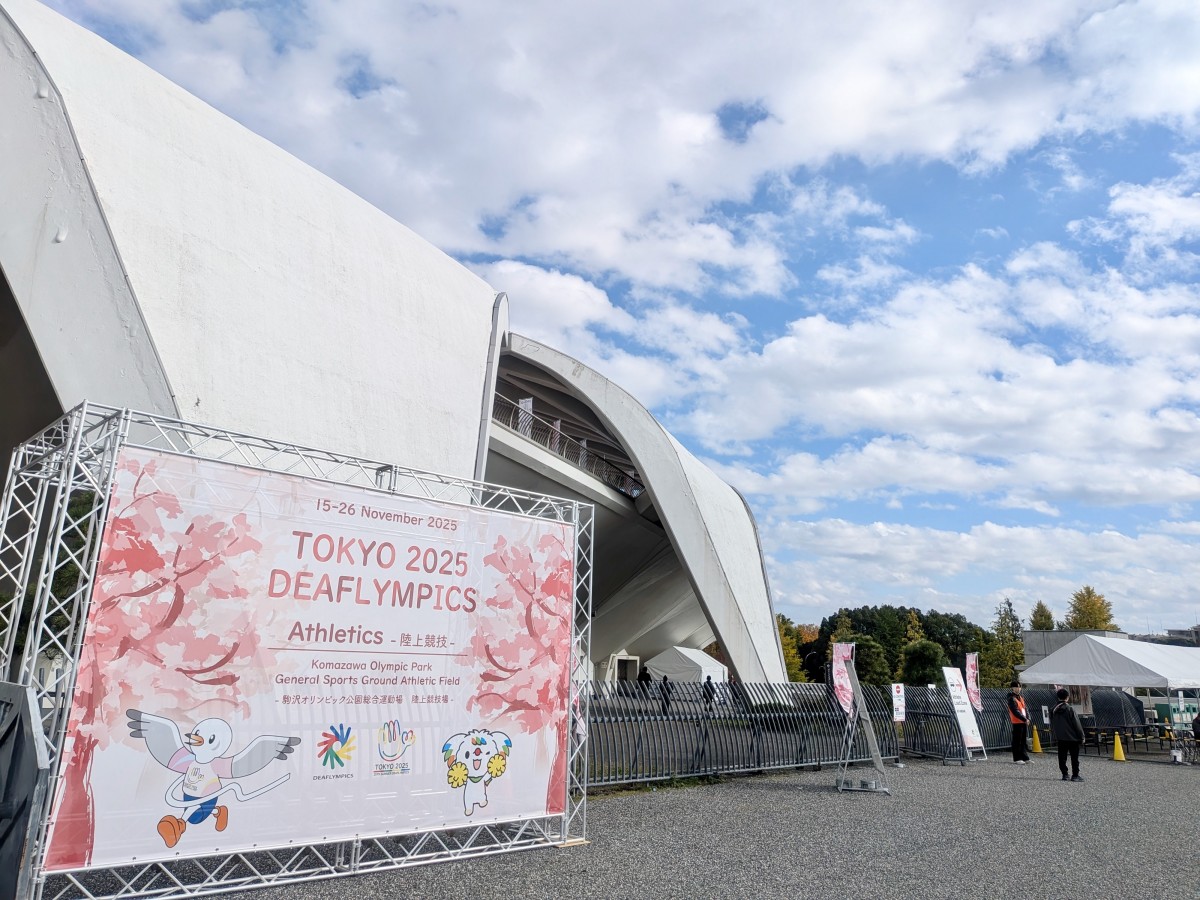 会場のひとつ駒沢公園。青空の下、白い屋根の競技場を背景に、デフリンピックの桜柄の巨大バナーが展示されている。