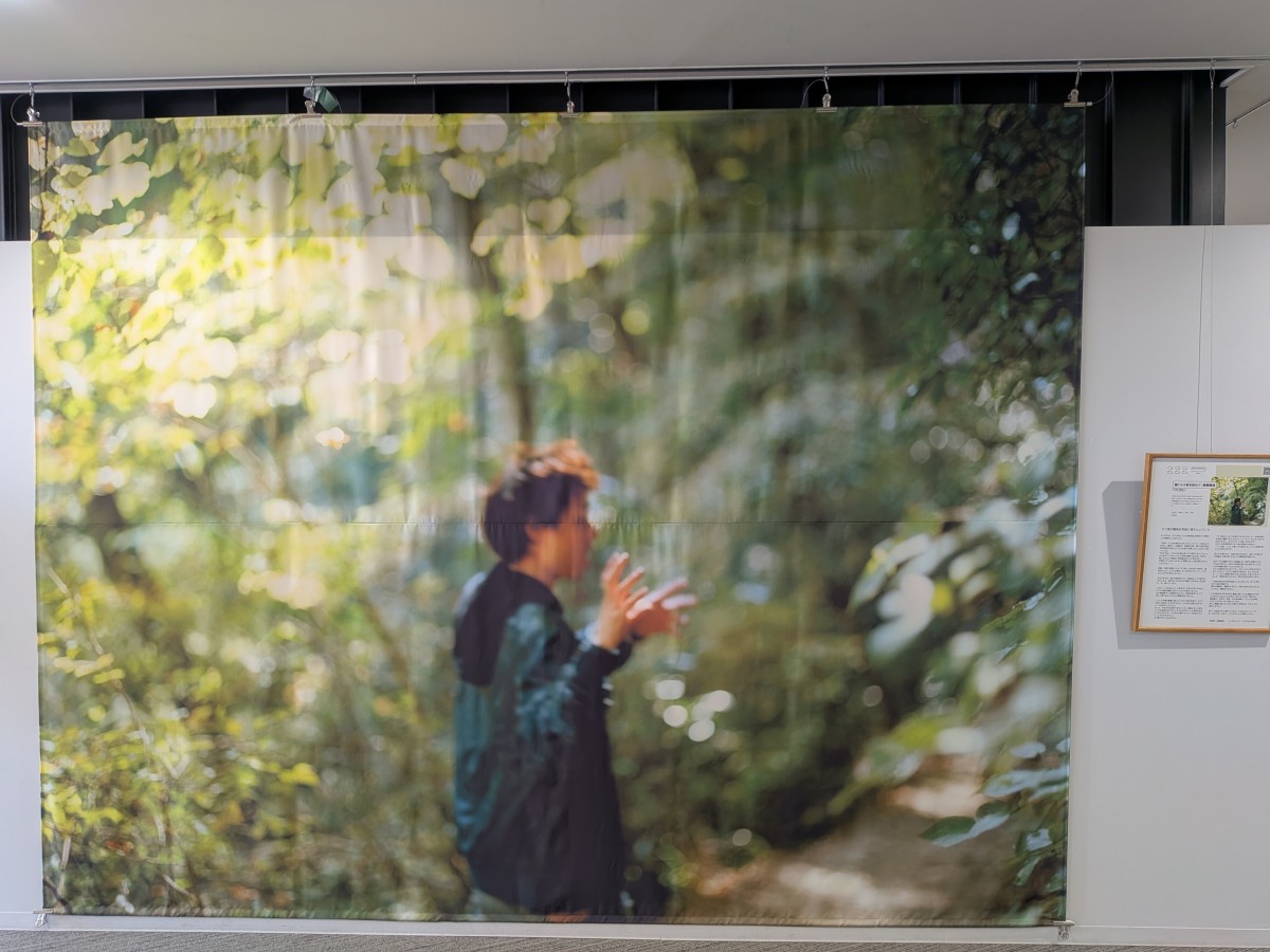 会場入口の大きな写真。深い木立のなかにパーカー姿の男性が佇んでいる。両手は指揮をするように振られようとしている