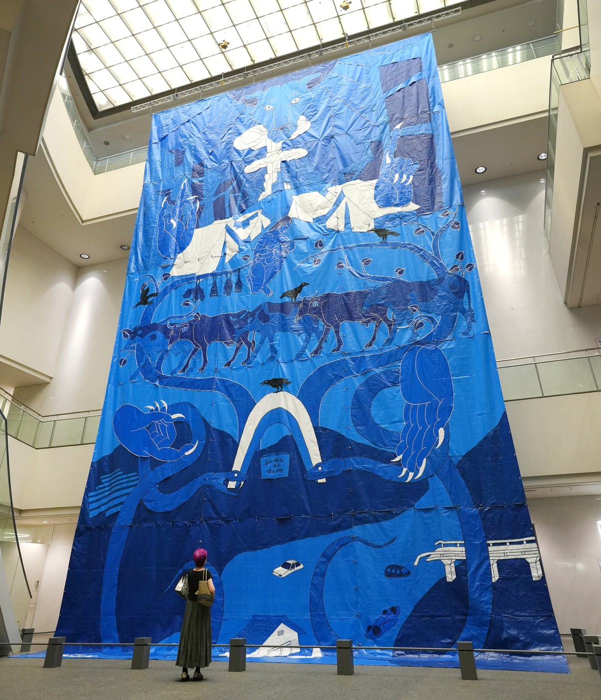 メイン会場の愛知芸術文化センターの吹き抜けに展示されている久保寛子さんの作品。ブルーシートをつなぎ合わせた絵柄には、きのこ雲から難民キャンプ、水害で流される家などがあり、隅々まで見入ってしまう=9月23日、関口撮影